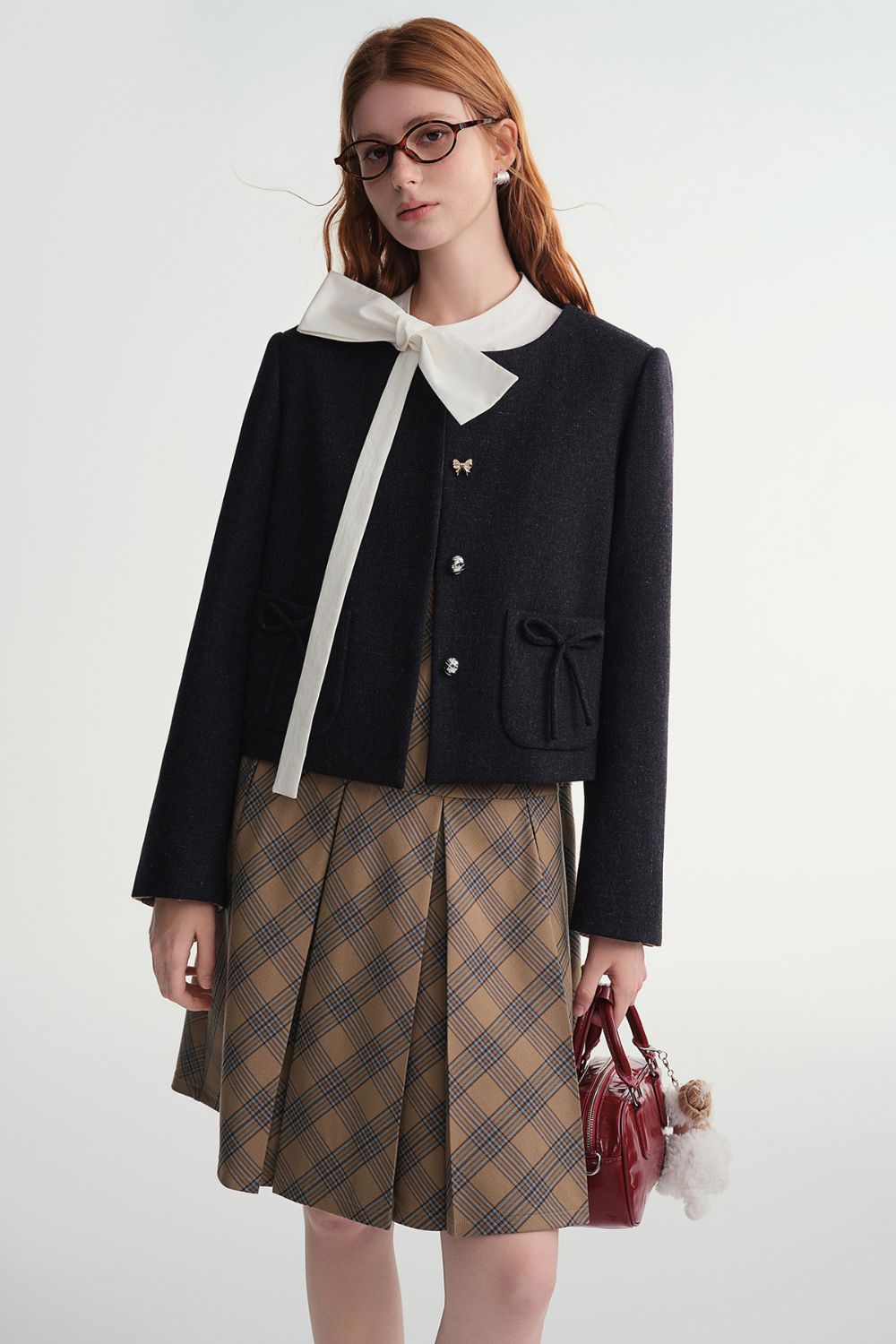 Model wearing a black cropped jacket over a white blouse with a bow tie and a brown plaid skirt