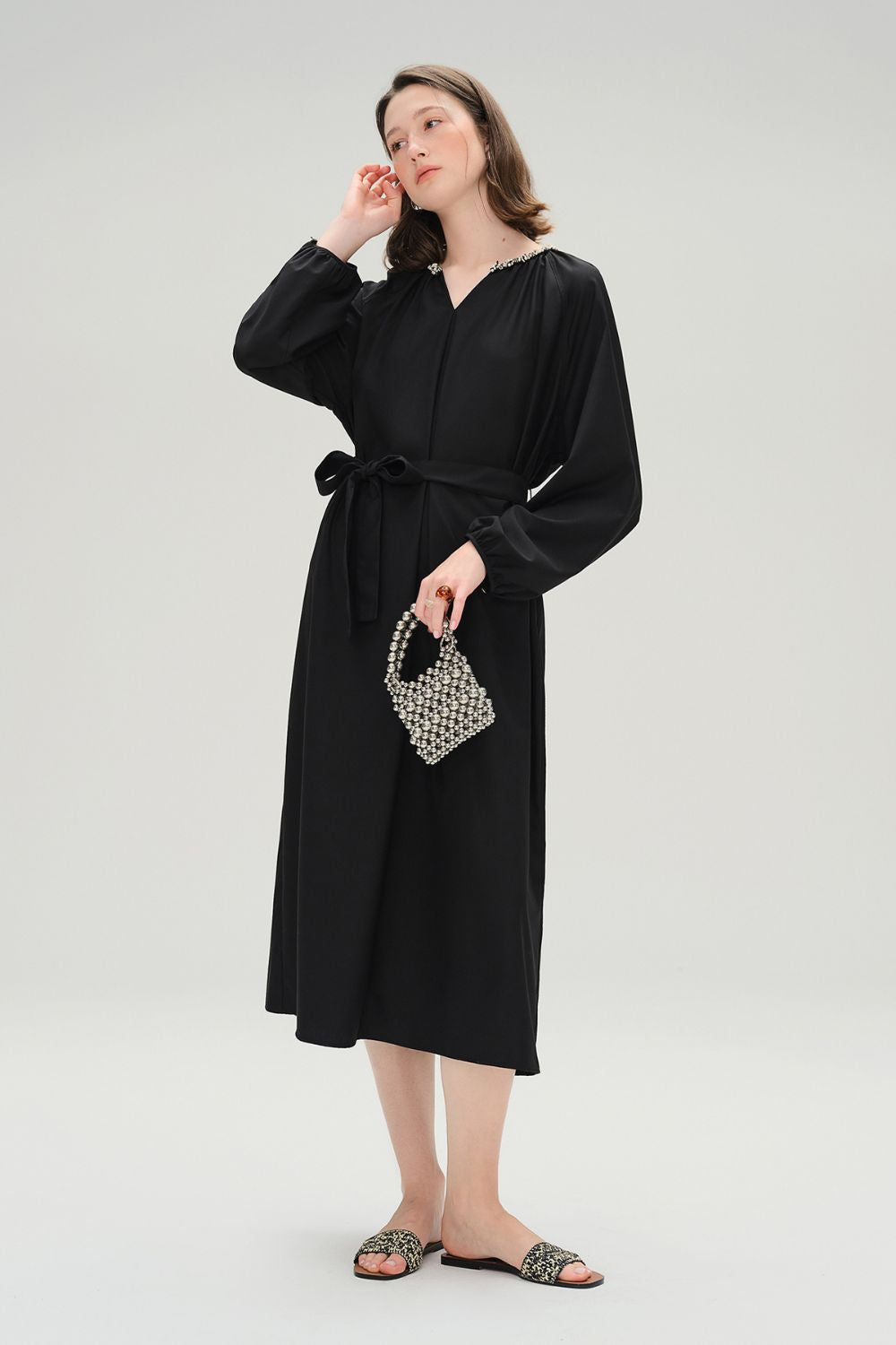 Model wearing black long-sleeve midi dress with tie waist, holding a beaded clutch