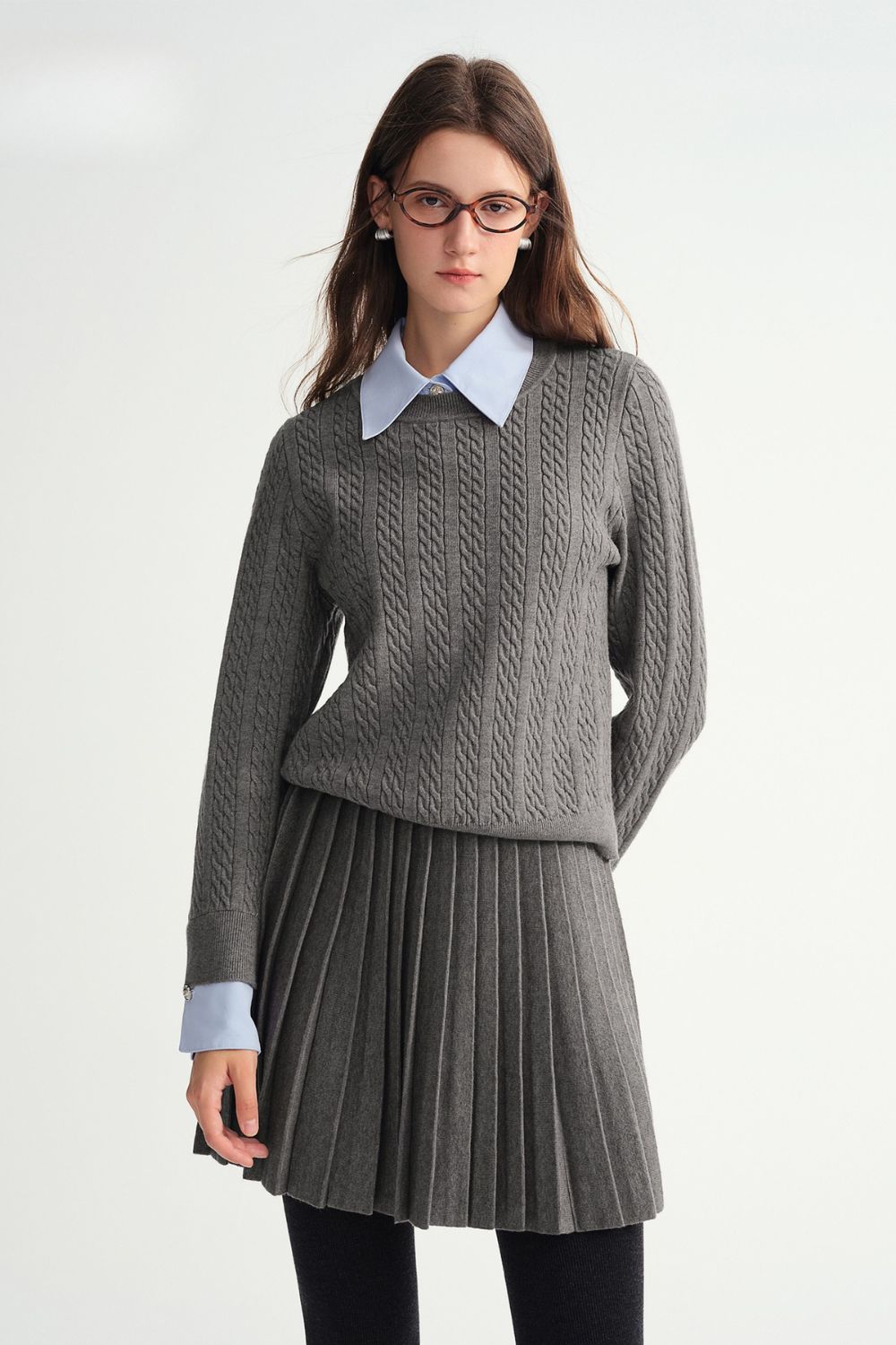 Model wearing gray cable-knit sweater layered over blue collared shirt with matching gray pleated skirt
