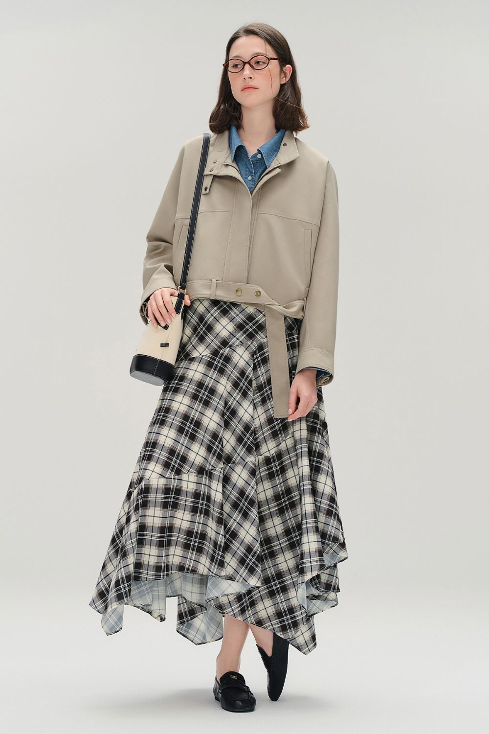 Model wearing a beige jacket and long plaid asymmetrical skirt with loafers