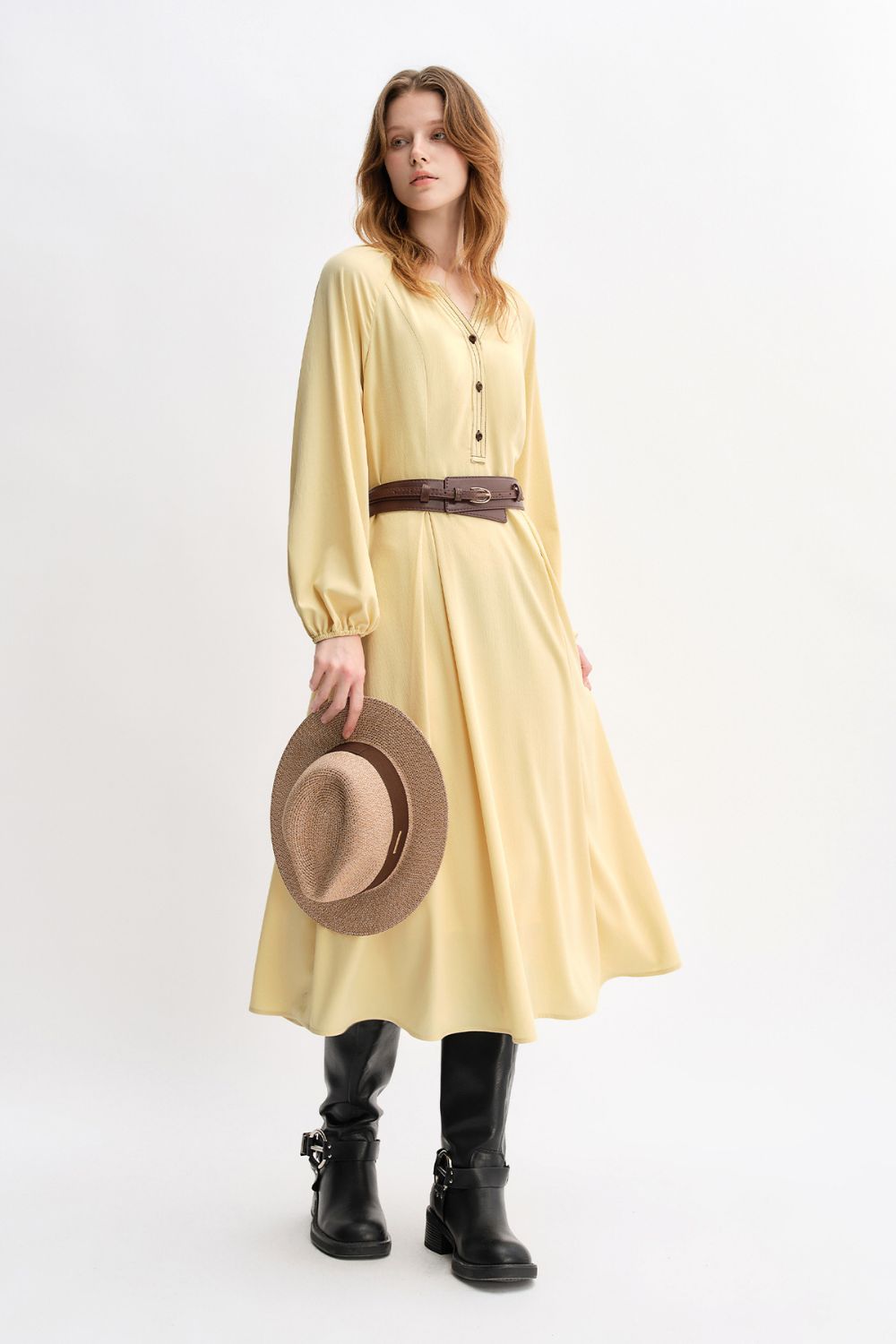 Model wearing a long yellow belted dress with long sleeves, holding a brown hat