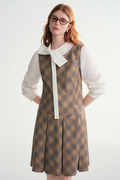 Model wearing sleeveless brown plaid A-line dress layered over a white blouse with a bow tie neckline