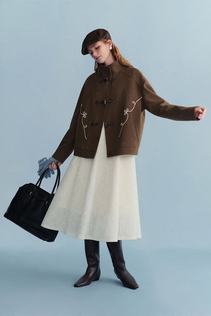 Model wearing a brown toggle jacket styled with a cream skirt and black boots, holding a handbag