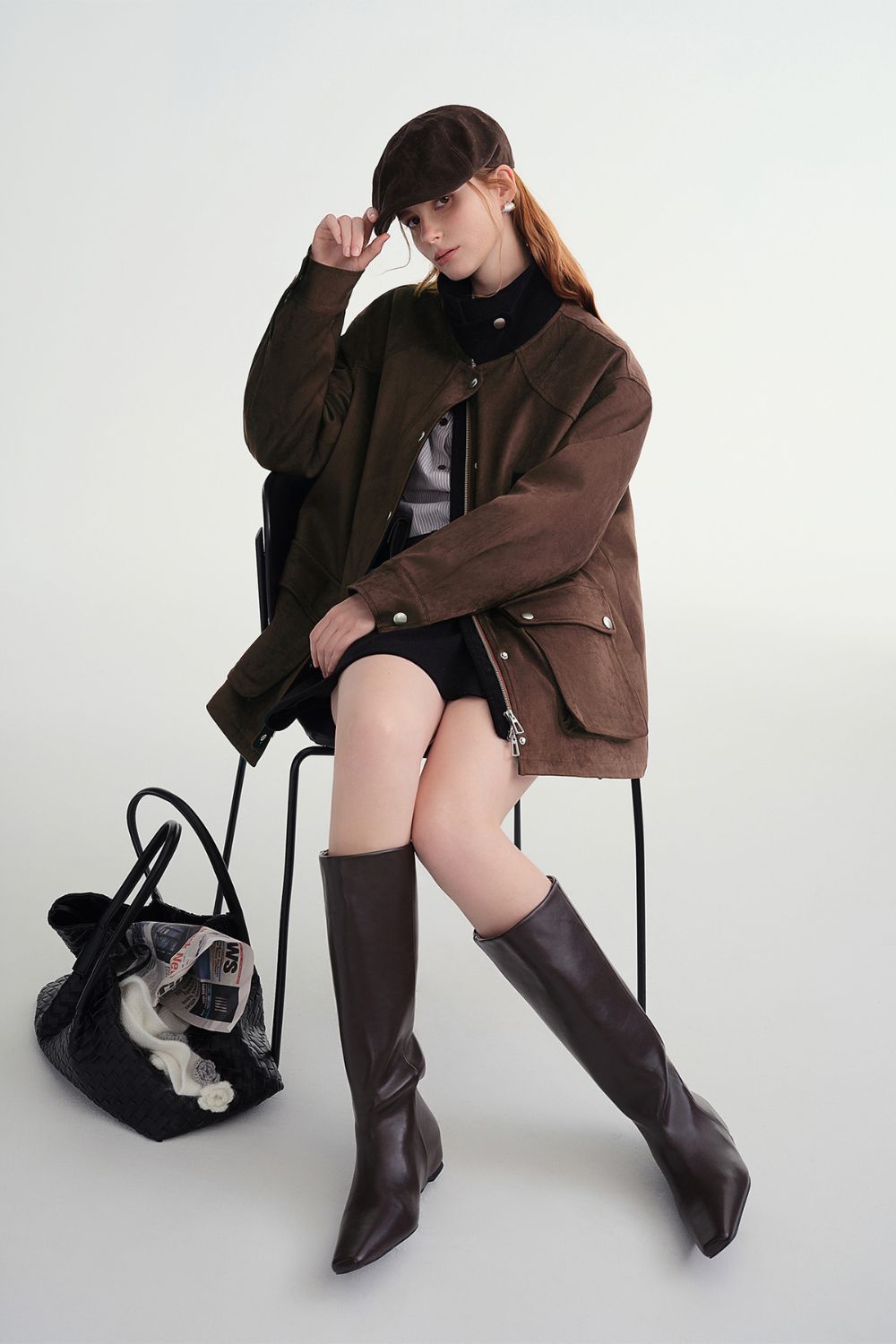 Model seated, wearing a brown button-up jacket with tall black boots and holding a bag