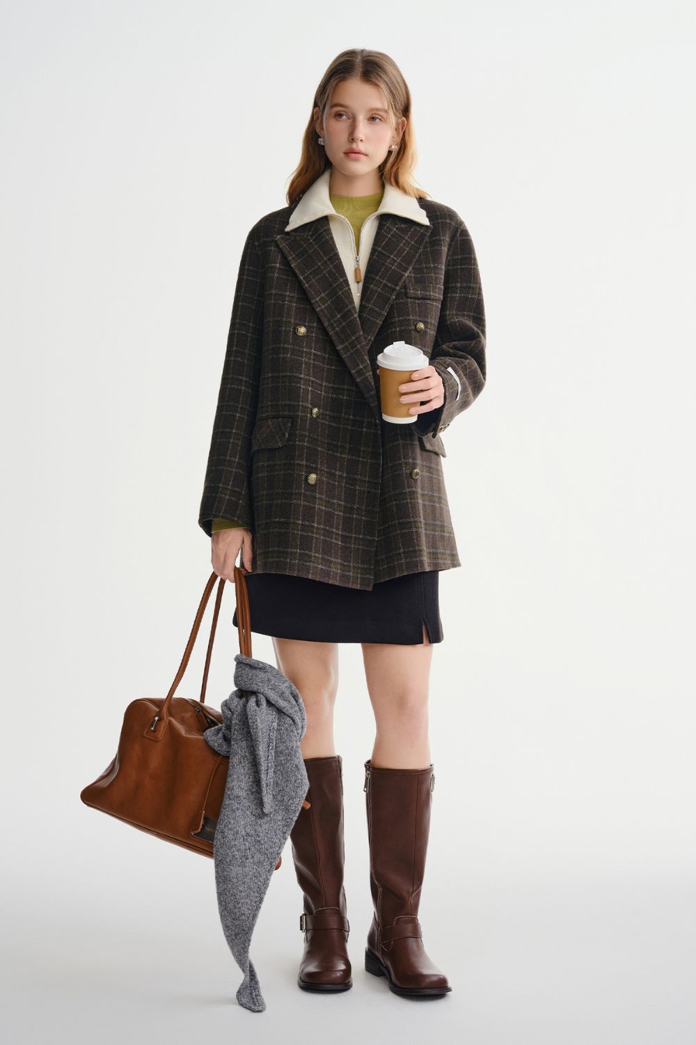 Model in plaid coat paired with a white blouse, mini skirt, and tall brown boots holding a handbag
