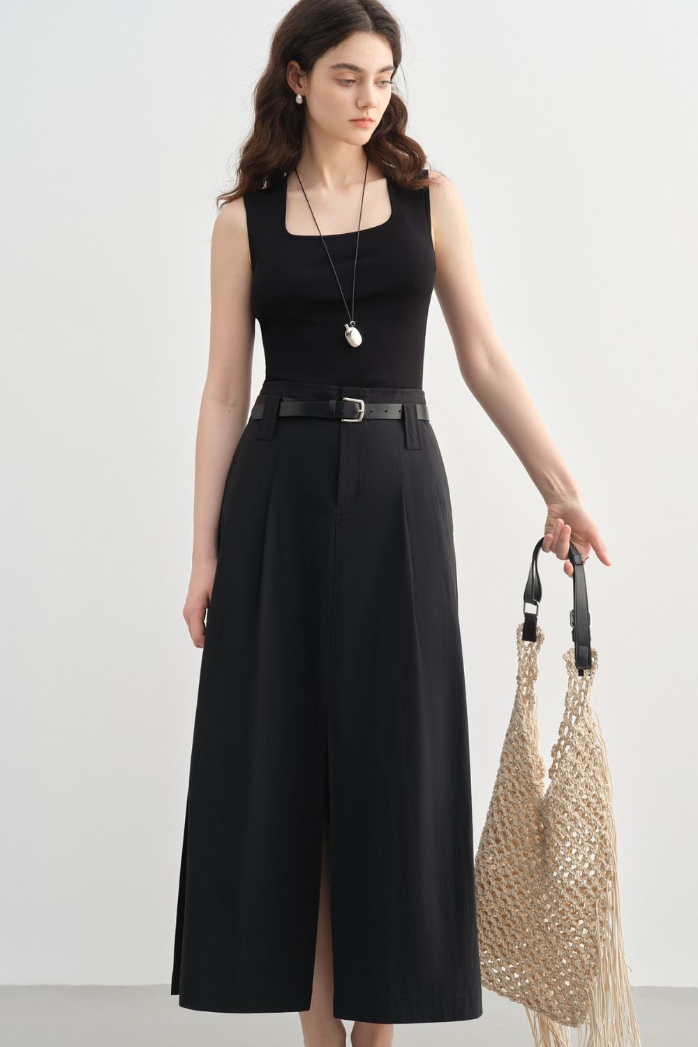 Model in a black sleeveless top and black midi skirt holding a woven bag